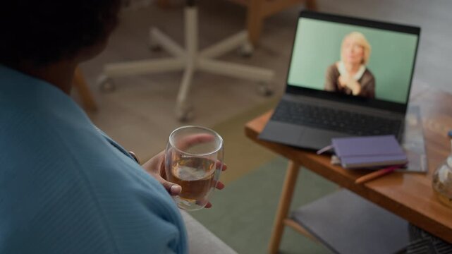 Rack focus of African American young woman video calling Caucasian girlfriend on wireless laptop and drinking tea sitting by table with notebooks and pregnancy test
