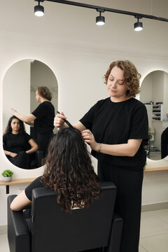 A female hairdresser using a styling tool on a client with natural curly hair. Professional beauty salon interior with mirrors and copy space. Concept of expert hair care and styling services.