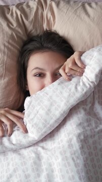 Young woman smiling and holding a soft duvet while lying in bed in the morning, embracing comfort and relaxation, enjoying a peaceful moment after waking up. Vertical shot