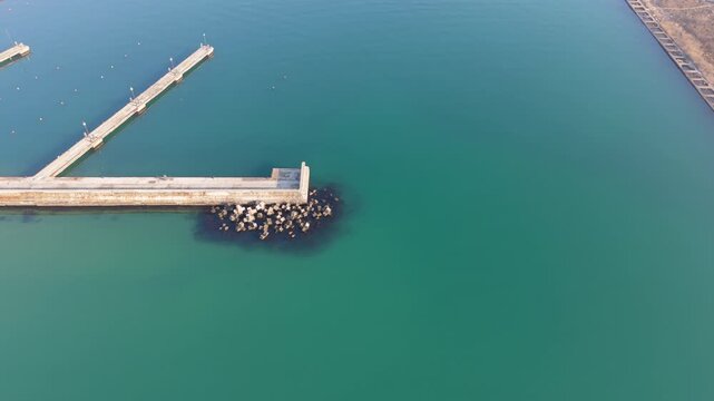 Aerial drone view of long concrete pier extending into calm turquoise sea with rocky breakwater protecting harbor entrance.