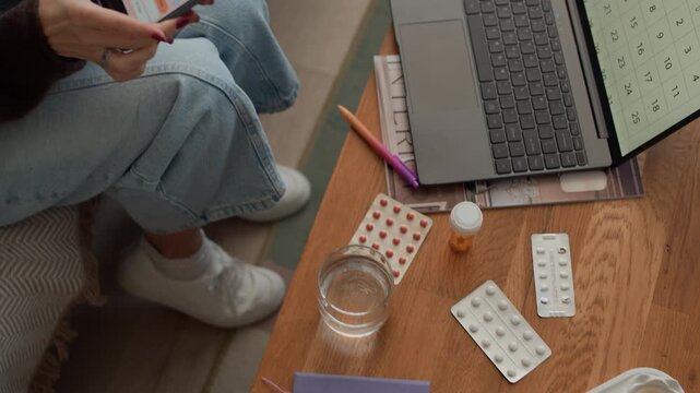 High angle handheld shot of middle-aged Caucasian woman tracking menstrual period calendar on smartphone while sitting by table with multiple pills and wireless laptop