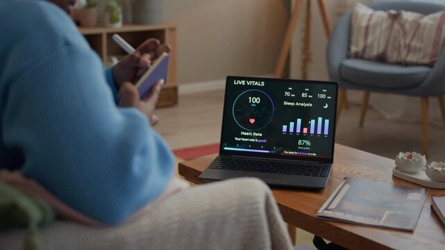 Cropped shot of young adult African American lady analyzing health indicators on screen of wireless laptop and taking notes while sitting on sofa in living room