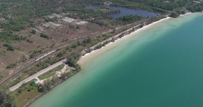 Forward aerial fuel tanker train on tracks parallel to a coastal road and white sandy beach with turquoise water, contrasting with a dark lake.