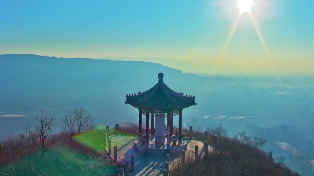 A serene high-angle shot of a classic Chinese gazebo on a hill overlooking a misty valley and distant city during a golden sunrise in North China.