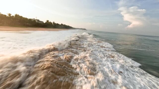 Aerial FPV view of the shoreline with foamy white waves crashing onto the sandy beach, creating a dynamic contrast with the dark blue ocean, Colombo, Western Province, Sri Lanka.