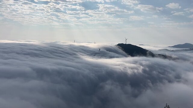 A stunning time-lapse shot of a thick sea of clouds flowing over mountain peaks with rotating wind turbines under a bright sky in northern China.