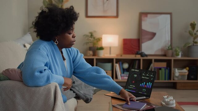 Long shot of young African American woman taking notes while sitting on couch and checking health progress on wireless laptop in living room