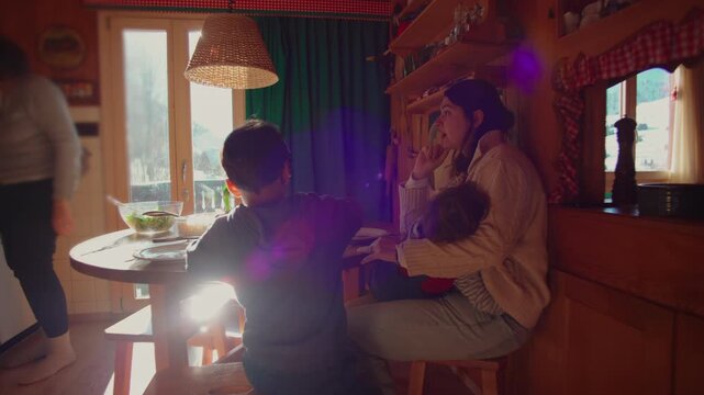 Mother sitting at wooden kitchen table holding toddler while older child eats nearby, warm family breakfast scene with sunlight through window in mountain home