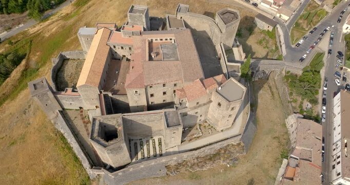 Aerial shot of the Castle of Melfi in Basilicata, Italy. View of a sprawling medieval stone castle with multiple towers and orange-tiled roofs. The fortification is situated on a golden, grassy hill.