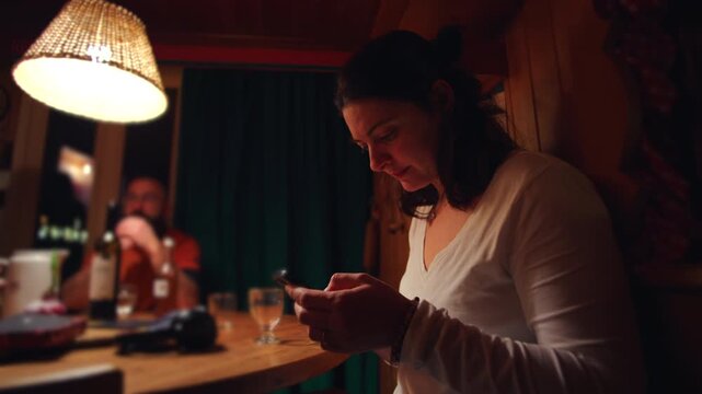 Woman checking smartphone at wooden dining table during family evening, partner blurred in background with wine bottle and glass, intimate home moment showing distraction and modern life