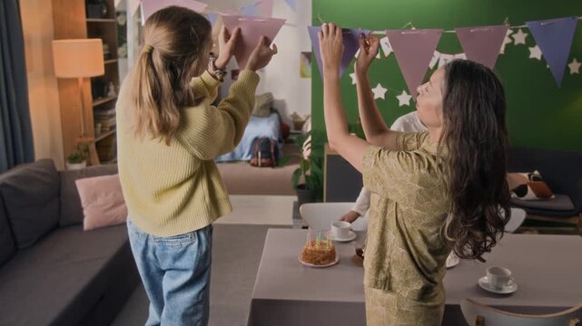 Mother and daughter adjusting festive flag garland and joyfully giving each other high-five while teenage boy setting table in background