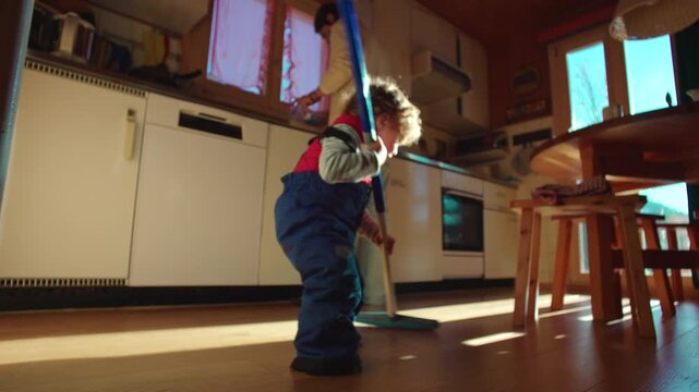Toddler sweeping wooden kitchen floor with mop while parent works in background, playful imitation of household chores in cozy family home