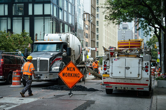 Construction workers in safety vests operate near a cement truck on an urban street. A bright orange men working sign warns drivers of active road repairs in the city.