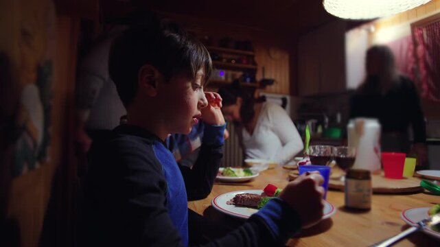 Young boy eating dinner at wooden table inside cozy alpine chalet, family gathered in warm kitchen during evening meal