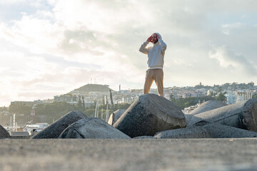 Energetic young man in white hoodie walking on concrete breakwaters against blue sunset sky,...