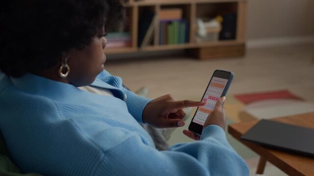 Arc shot of female young Black user checking menstrual cycle on her smartphone while sitting on couch in cozy living room