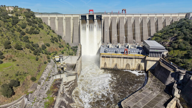 Aerial view of the Alcantara Dam unleashing powerful torrents against a backdrop of rugged terrain under a clear sky, Alcantara, Extremadura, Spain.