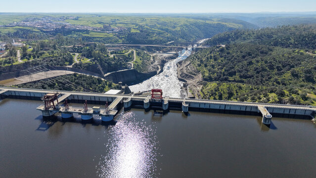 Aerial view of the Alcantara Dam releasing water into the Tagus River, framed by verdant hills and the distant town, Alcantara, Extremadura, Spain.
