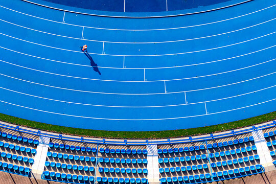Aerial view of a solitary figure graces the vibrant blue running track within the stadium, a striking contrast against the rows of empty blue seats, Warsaw, Masovian Voivodeship, Poland.