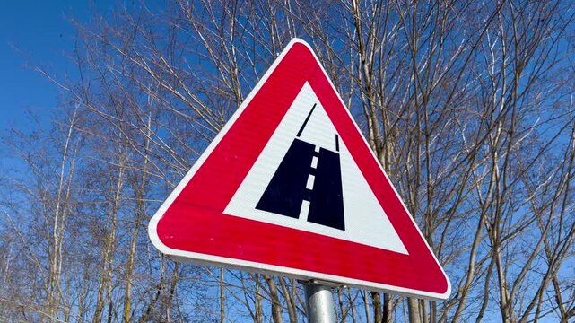 Road surface change warning sign indicating asphalt to gravel transition on rural roadside with bare trees and blue sky in late winter countryside landscape Latgale Latvia
