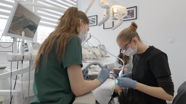 Female dentist and medical assistant use dental drill and saliva ejector suction tube to treat caries cavity in teenage boy patient mouth during family stomatology clinic appointment.