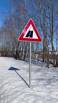 Road surface change warning sign standing in snowy roadside landscape with bare trees and blue sky at end of winter, rural transportation symbol in Latgale countryside