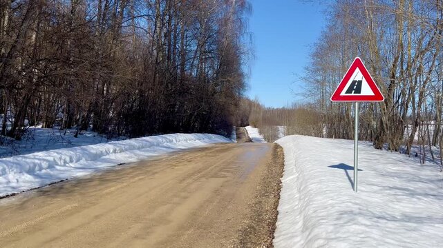 Road surface change warning sign beside rural gravel road with snowbanks and forest in late winter countryside near Razna Lake, seasonal Baltic landscape