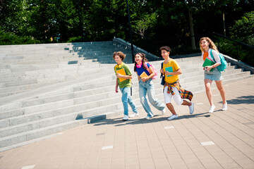 Fototapeta premium Group of cheerful young students walking outdoors on a sunny day holding book and backpacks near urban stairway