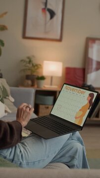 Vertical shot of cheerful middle-aged Caucasian woman calling on mobile phone while checking pregnancy calendar on laptop, sitting on couch in modern living room