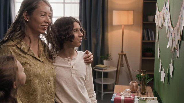 Mother adjusting paper star and stepping back to examine festive garland on wall with two positive gen alpha kids, then giving it thumbs-up