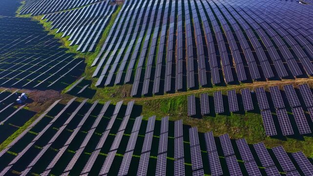 Rows of solar panels stretch across a vast field, collecting energy under the bright sun. The landscape is shaped by green patches and paths between the panels.