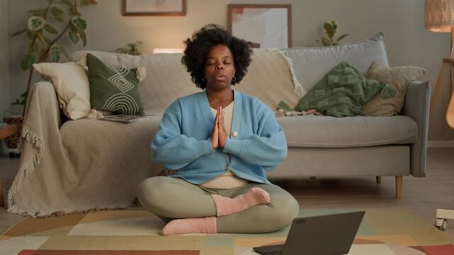 Long shot of Black young adult woman turning on meditation app on wireless laptop while sitting on floor in lotus pose in modern living room