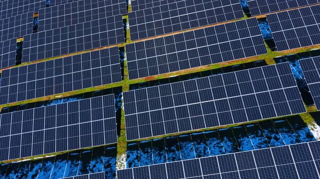 Angled aerial shot of solar panel arrays with snow. Perspective view of photovoltaic panels installed on ground with patches of melting snow and green grass.