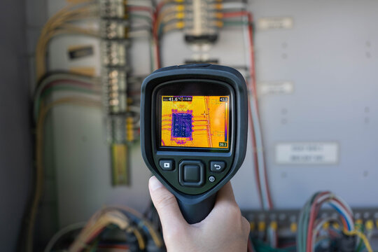 Thermal camera infrared inspection monitoring of control panel site; technician hand scans wiring to detect overheating prevent failures, support predictive maintenance and safety.