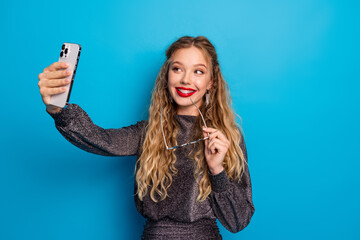 Fototapeta premium Young woman in a shiny dress takes a selfie against a blue background wearing glasses with a glamorous smile for a festive stylish photoshoot