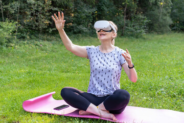 Fototapeta premium Excited Mature Woman Playing Interactive VR Game on Yoga Mat Outdoors. Lady Using Virtual Reality Headset and Smartphone for Gaming in Park