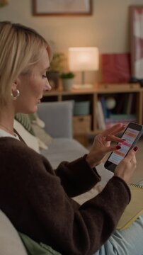 Vertical shot of middle-aged Caucasian woman tracking menstrual cycle in app on smartphone and taking notes while sitting on sofa in living room