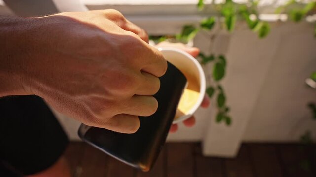 Milk Pouring Into Fresh Coffee in White Cup Creating Latte Art Pattern While Held in Hand on Wooden Terrace With Green Plants Background, Close Up View Showing Coffee Preparation