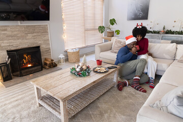 Naklejka premium African American couple lounging in living room on sofa by fireplace wearing Santa hats, copy space