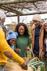 Diverse multigenerational family harvesting, sorting leeks from crate under pergola in knit hats