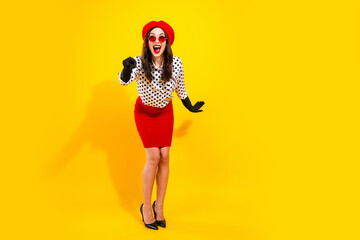 Obraz premium Happy young woman in red beret and pencil skirt wearing polka dot blouse, black gloves and red sunglasses posing and pointing on a bright yellow background in retro fashion style