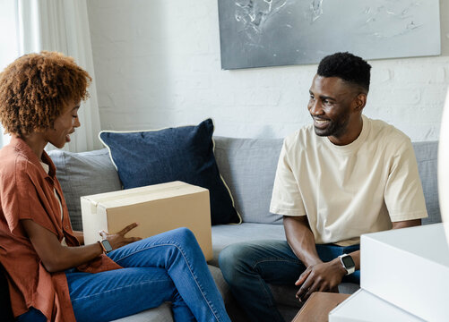 African American couple holding cardboard box on lap, smiling at home on gray sofa