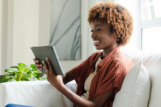 African American woman sitting on sofa at home wearing rust shirt holding tablet near potted plant
