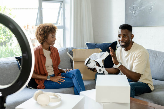 Diverse couple sitting on gray sofa at home holding white VR headset and smiling