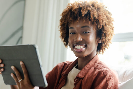 African American woman wearing rust shirt sitting on light sofa by bright window holding tablet