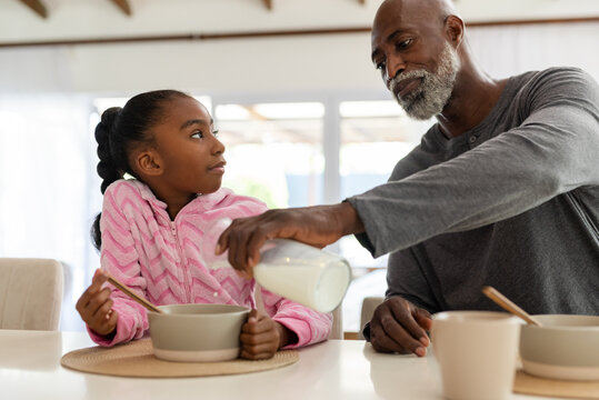 African American grandpa pouring milk from bottle into bowl as granddaughter holding spoon at table