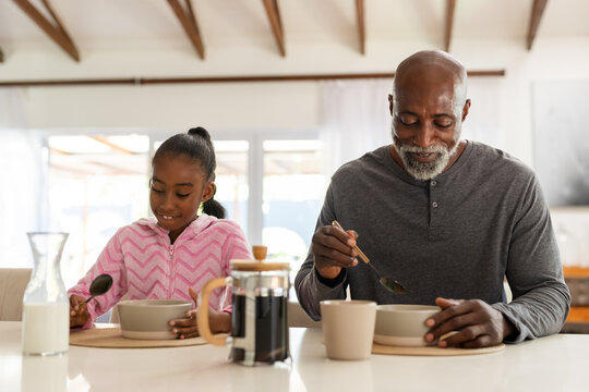 African American mature man and youth girl eating at home counter with French press, pink hoodie