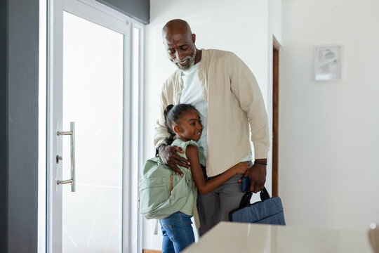African American senior father hugging child daughter in home foyer holding briefcase and backpack
