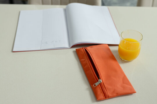 Orange fabric pencil case resting on cream tabletop near window, notebook and glass of juice nearby