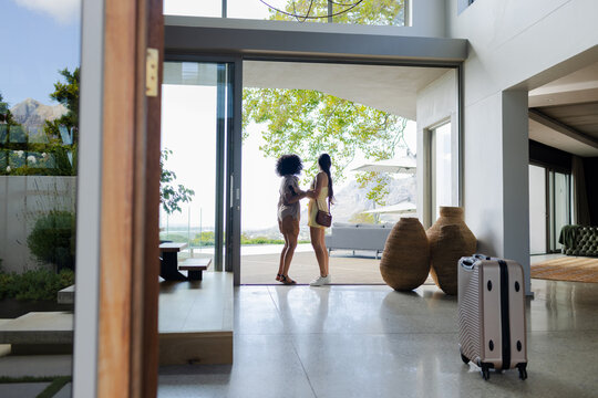 Diverse female friends wearing summer outfits, holding hands near sliding glass doors with suitcase